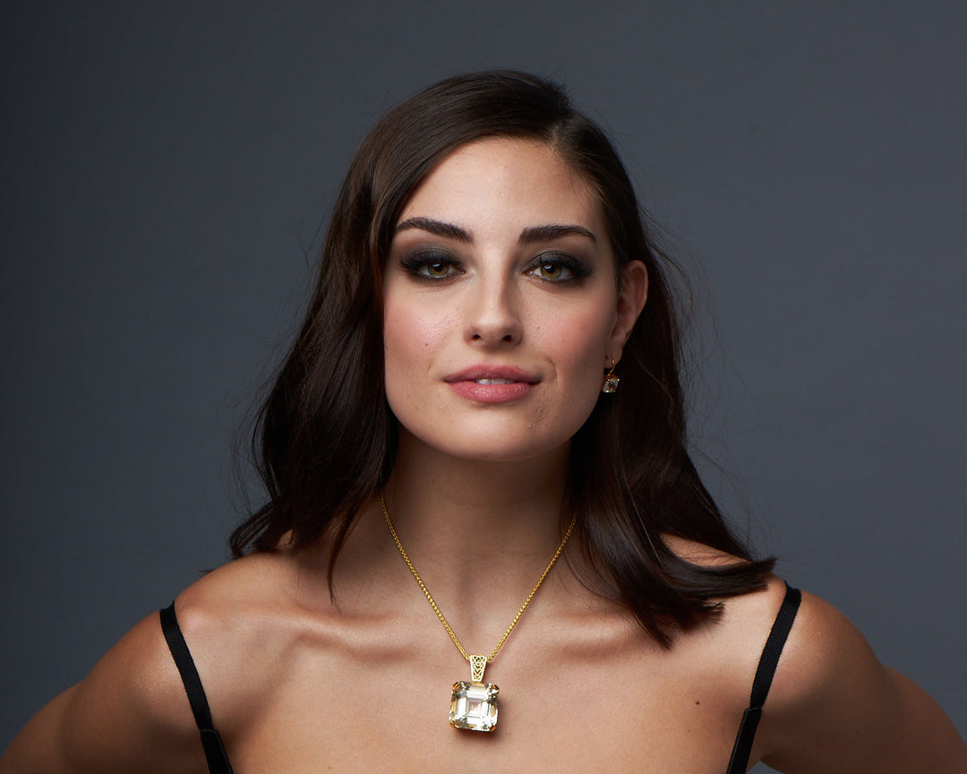 A woman wearing a gold necklace with a large crystal gemstone pendant and matching earrings, symbolizing calm and balance. The crystal gemstone pendant is often associated with reducing anxiety and stress.