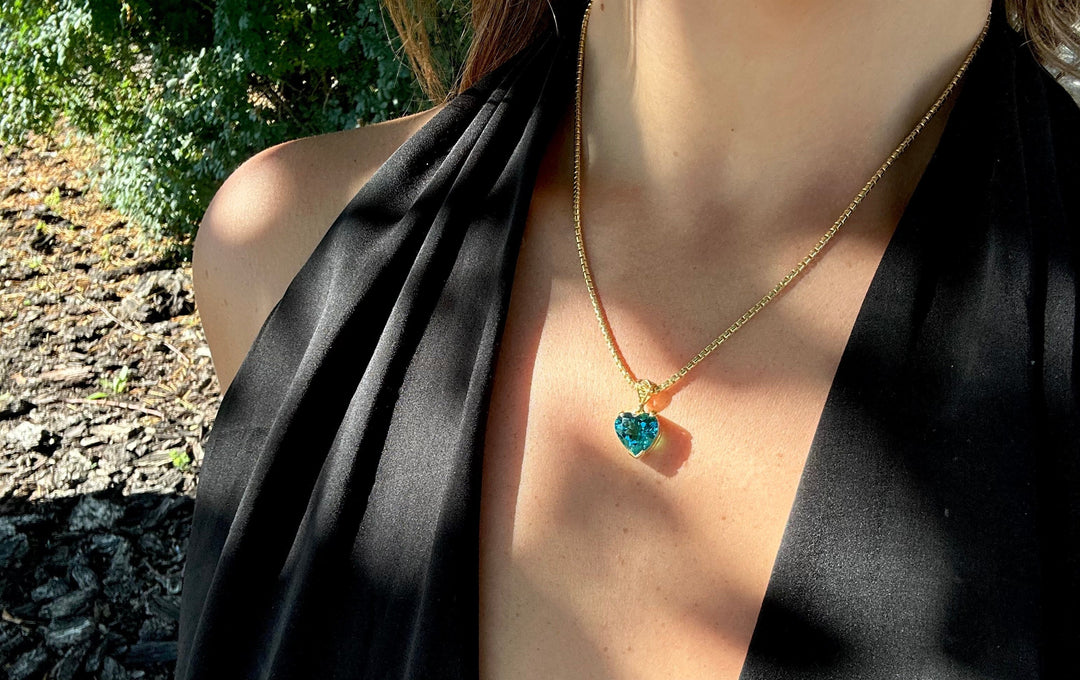 Close-up of a woman wearing a gold chain with a blue heart crystal gemstone pendant, symbolizing healing properties, self love, and protection against negative energy.